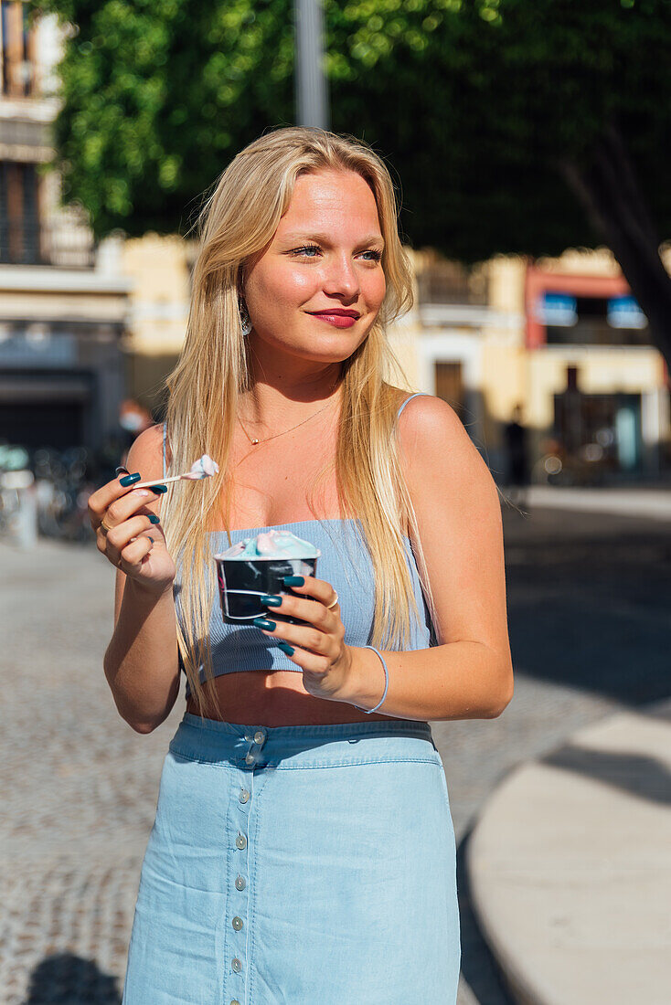 Beautiful blonde young female eating cold tasty ice cream while standing in city street on sunny day in summer