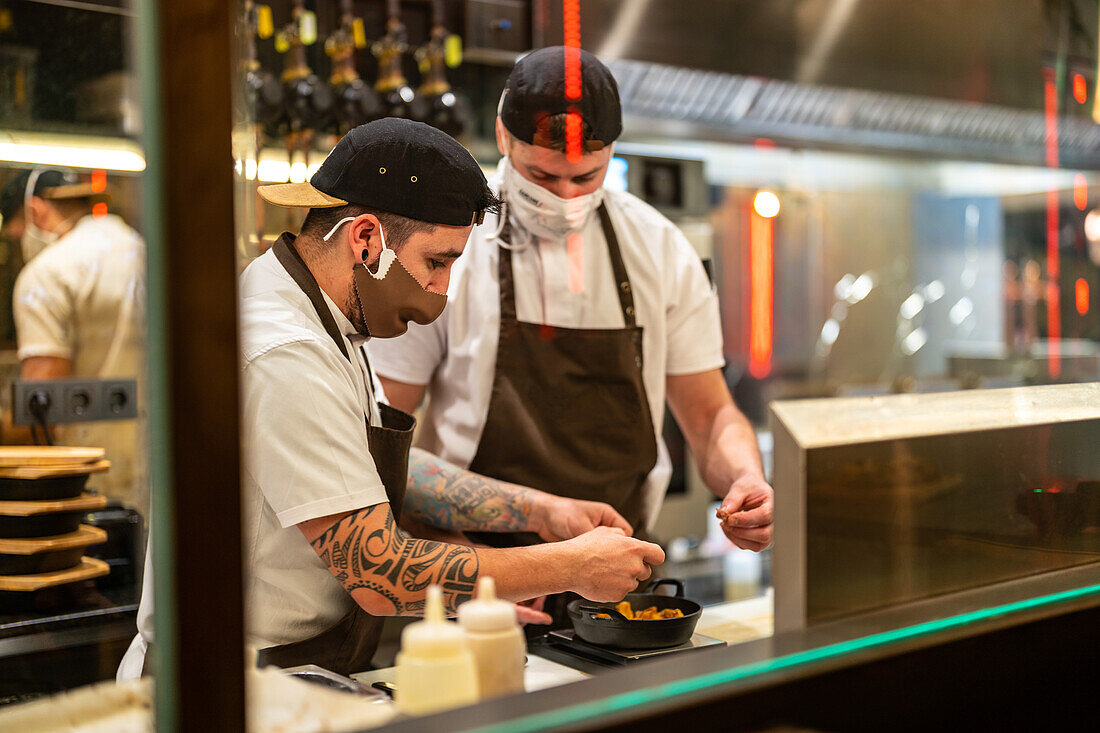 Männliche Köche in eleganter Uniform und mit Schutzmasken zur Verhinderung der Ausbreitung von COVID 19 bei der Zubereitung einer Mahlzeit in der Küche eines Restaurants (durch Glas)