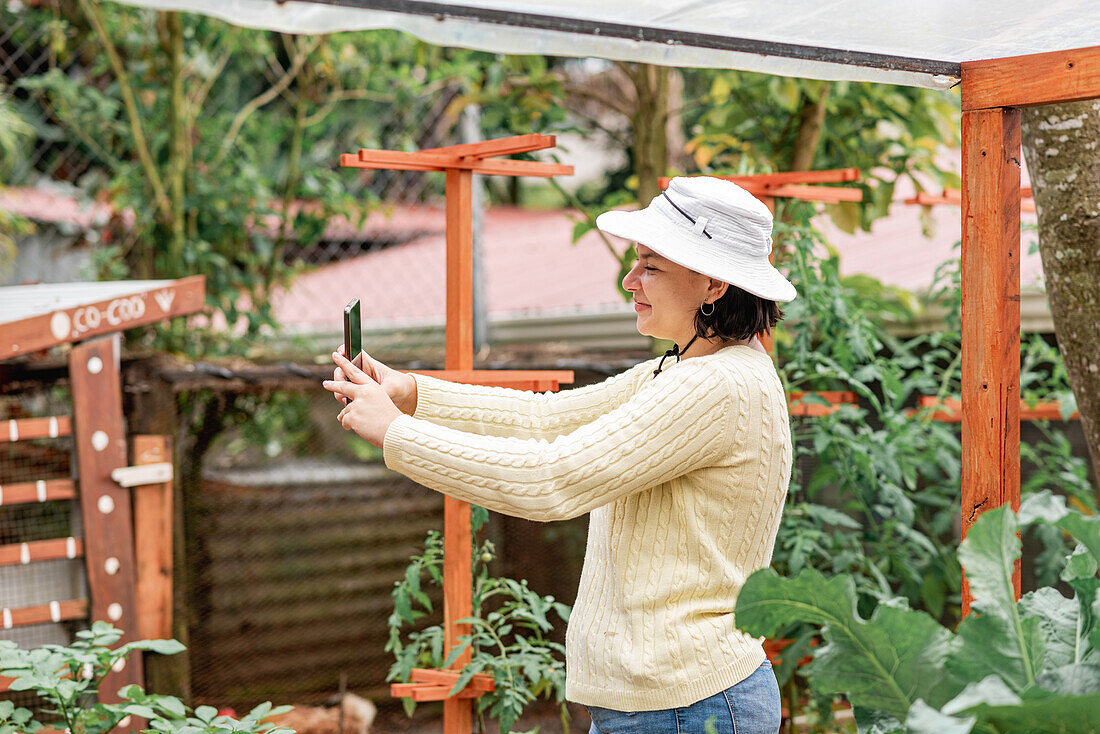 Seitenansicht einer lächelnden Gärtnerin, die zwischen Pflanzen in Gartenbeeten steht und ein Selbstporträt mit dem Handy macht