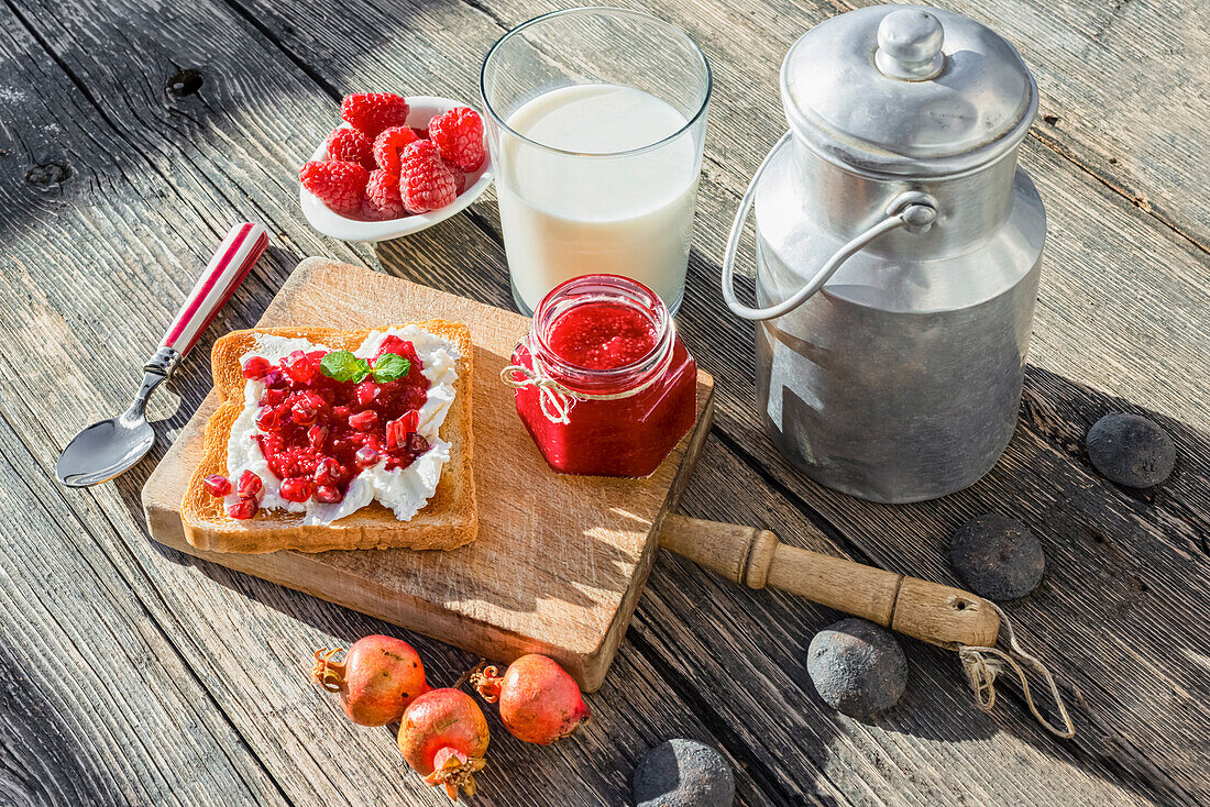 From above of yummy homemade toast with jam and mini pomegranates places near plate with raspberry and a large glass of milk next to a brass pitcher on wooden table
