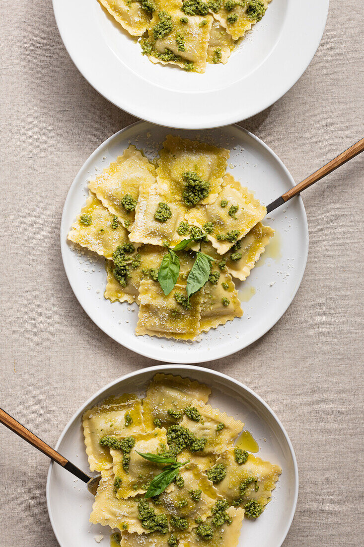 Draufsicht auf appetitlich gekochte Ravioli mit grüner Soße und Kräutern auf weißen Tellern mit Gabeln auf dem Tisch