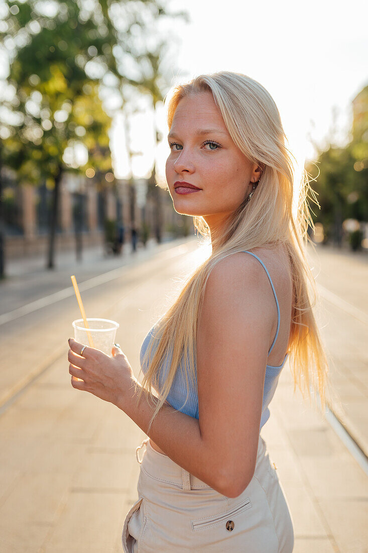 Seitenansicht einer Frau mit kalter Limonade in einem Plastikbecher auf der Straße im Sommer, die in die Kamera schaut