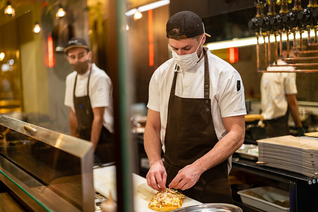 Männlicher Koch in Uniform und Maske zur Vorbeugung gegen das Coronavirus, der in einem Restaurant Käse auf gebackenes Fladenbrot gibt