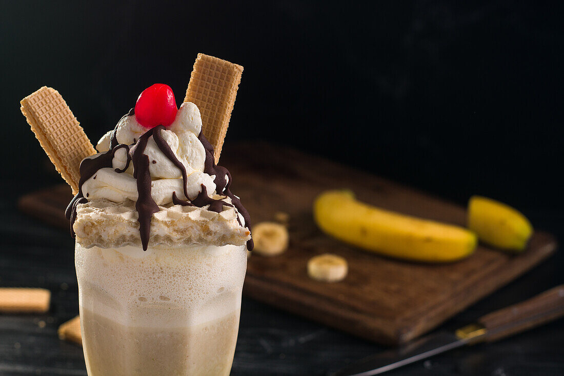 Glas süßer Bananenmilchshake, garniert mit Schlagsahne, Waffeln und Kirsche mit Schokolade obenauf