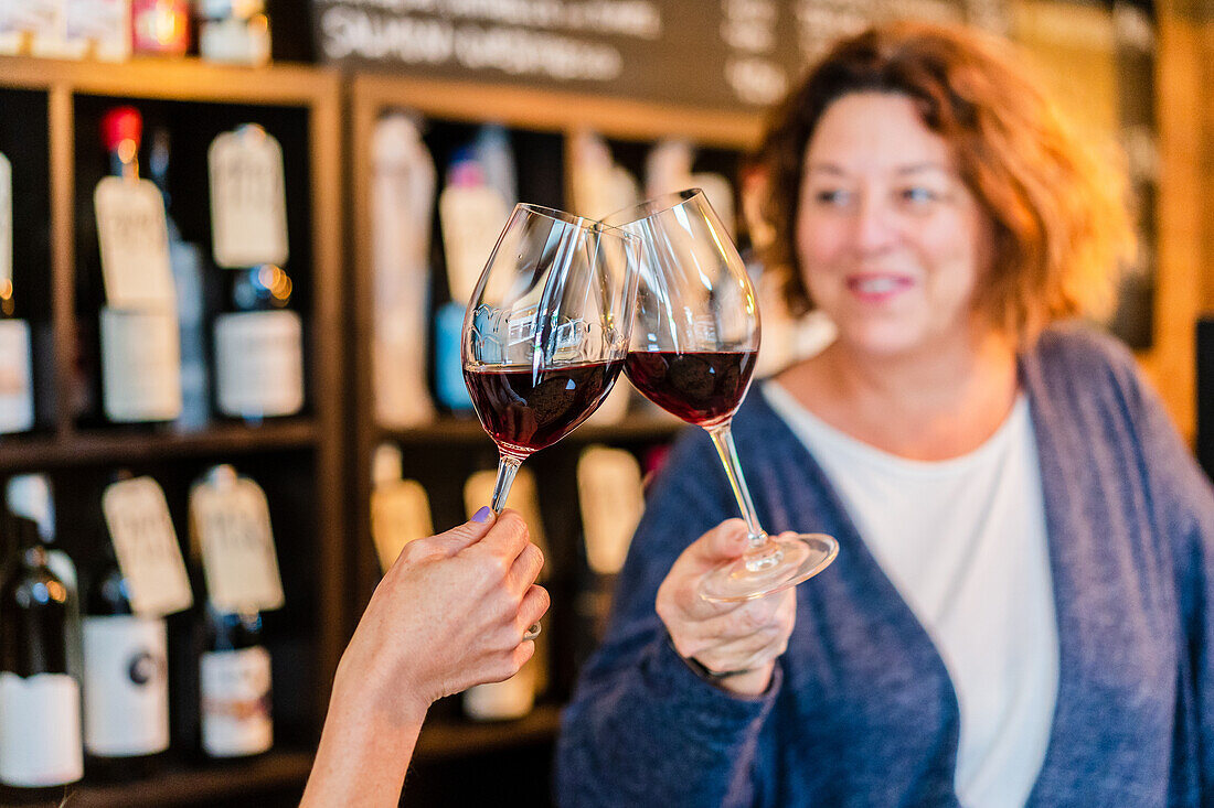 Anonyme Frau mit Maniküre stößt mit Gläsern mit aromatischem Rotwein an, während ein unscharfer, fröhlicher Freund vor einem Regal mit Weinflaschen in einem Restaurant steht