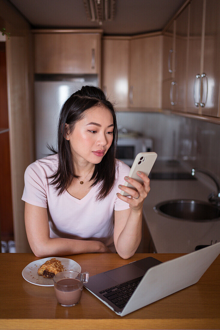 Positive asiatische Frau schreibt Textnachrichten auf dem Handy, während sie am Tisch mit Netbook und Essen mit heißem Getränk in der Küche sitzt