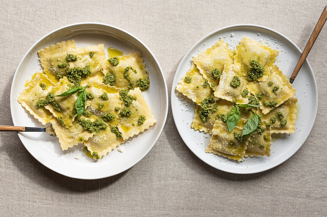 Draufsicht auf appetitlich gekochte Ravioli mit grüner Soße und Kräutern auf weißen Tellern mit Gabeln auf dem Tisch