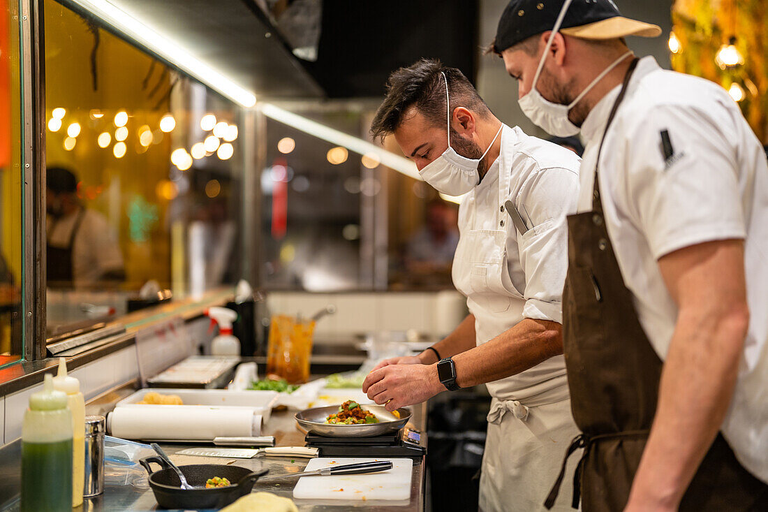 Seitenansicht eines professionellen Kochs mit Maske, der mit einem Kollegen in einer Restaurantküche während einer Coronavirus-Pandemie einen Teller mit einem köstlichen Gericht serviert