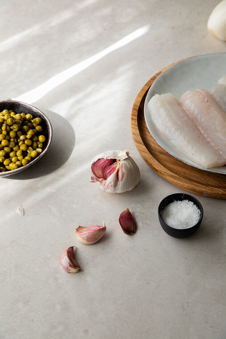 Frische Knoblauchzehen und Salz auf dem Tisch neben Seehechtfilet und einer Schüssel mit Erbsen bei der Essenszubereitung in der Küche von oben gesehen