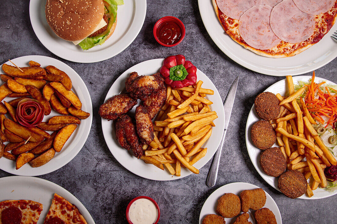 Draufsicht auf Teller mit verschiedenen Junkfood-Gerichten, die während des Mittagessens auf einem grauen Tisch neben Schüsseln mit Soßen stehen