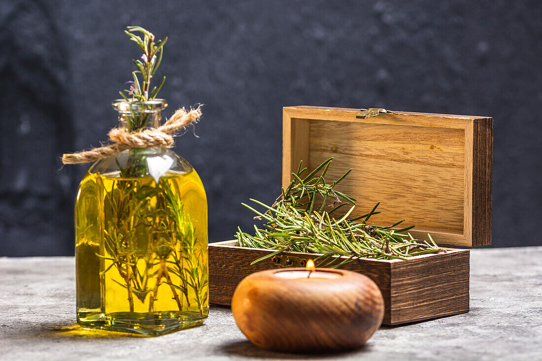 Glasflasche mit ätherischem Öl, Rosmarinzweigen und brennender Bio-Holzkerze auf grauem Tisch