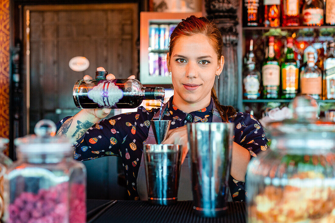 Zufriedene Barkeeperin gießt Alkohol in einen Metallshaker und bereitet einen erfrischenden Cocktail an der Theke einer Bar zu