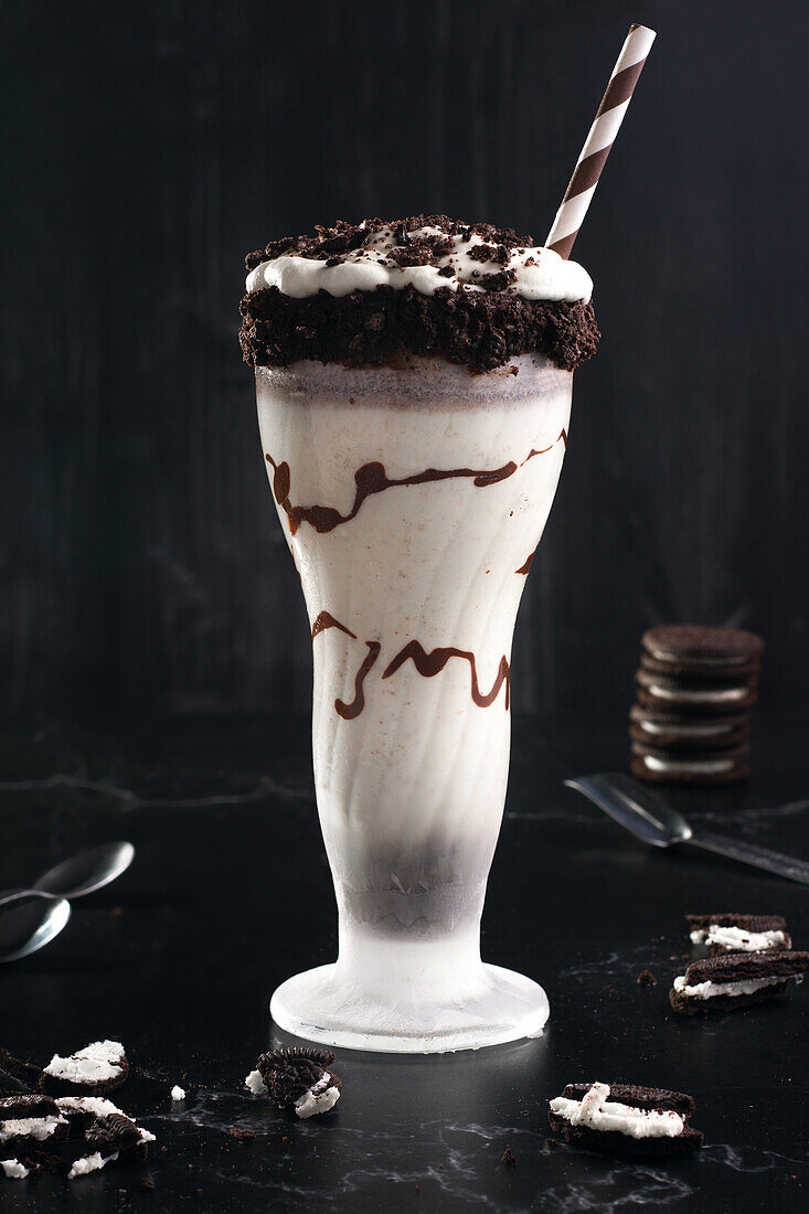 Tasty milkshake with crushed biscuits and straw in glass with chocolate sauce
