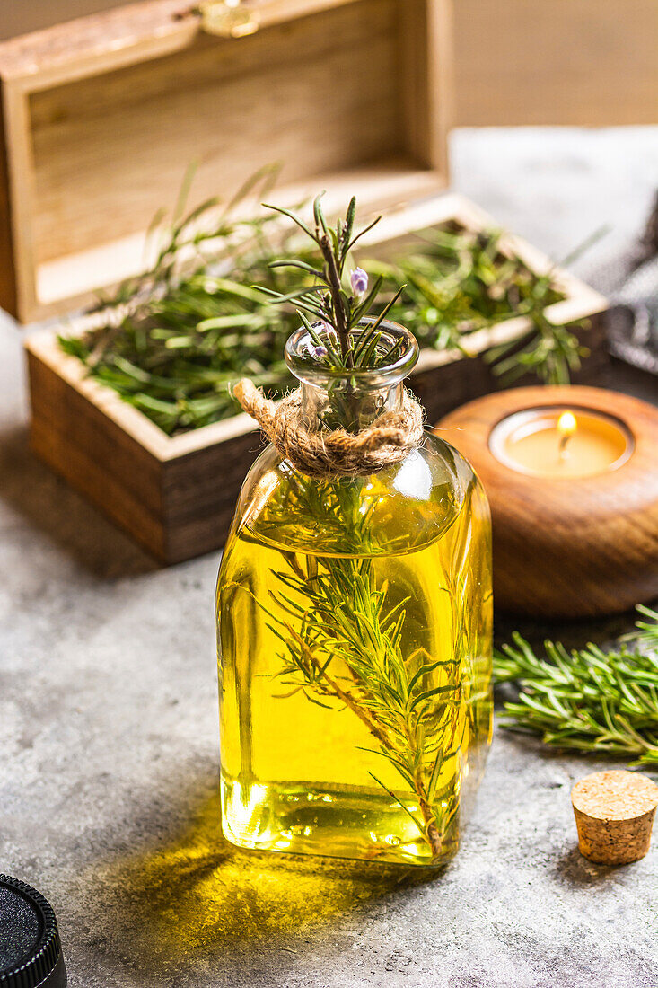 Glasflasche mit ätherischem Öl, Rosmarinzweigen und brennender Bio-Holzkerze auf grauem Tisch