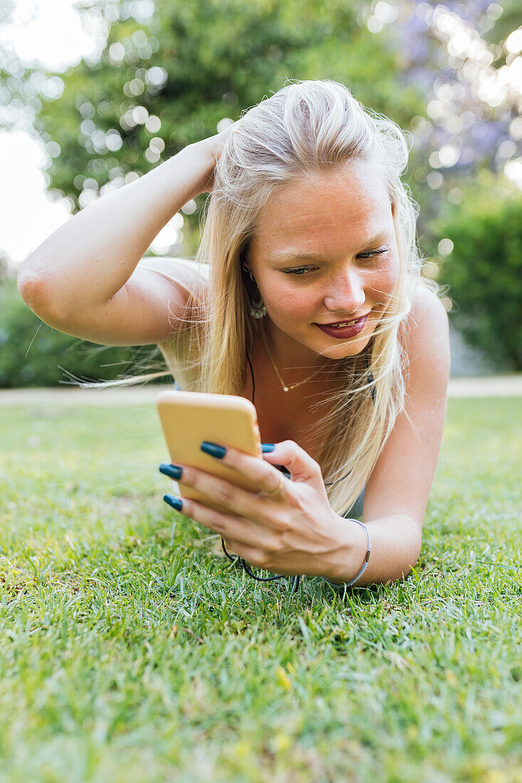 Lächelnde, charmante Frau, die im Sommer im Gras im Park liegt und mit Kopfhörern Musik hört