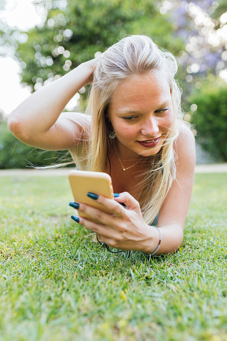 Smiling charming female lying on grass in park and listening to music in headphones in summer