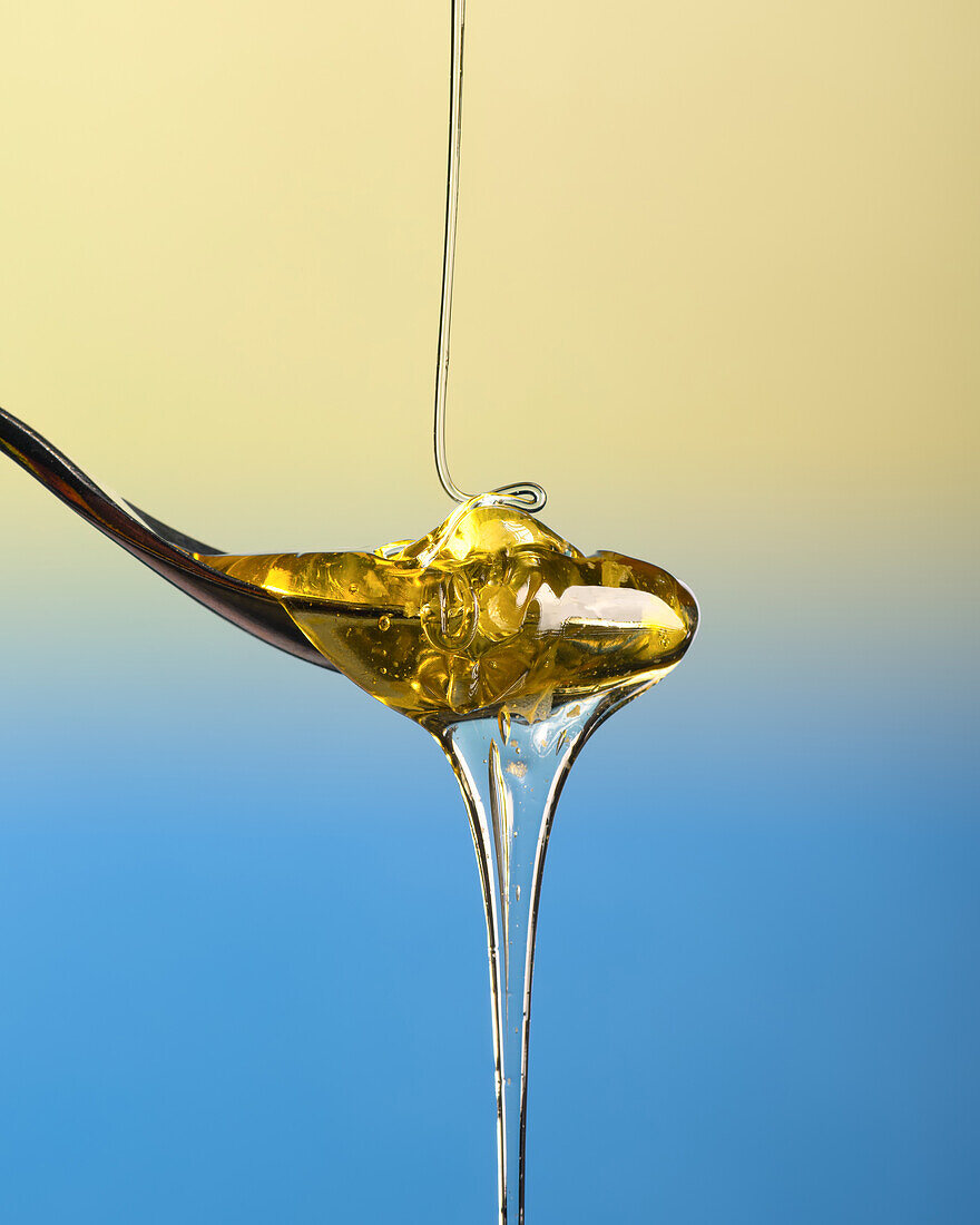 Stream of clear honey pouring onto a teaspoon and overflowing, isolated, on a plain blue and yellow gradient background