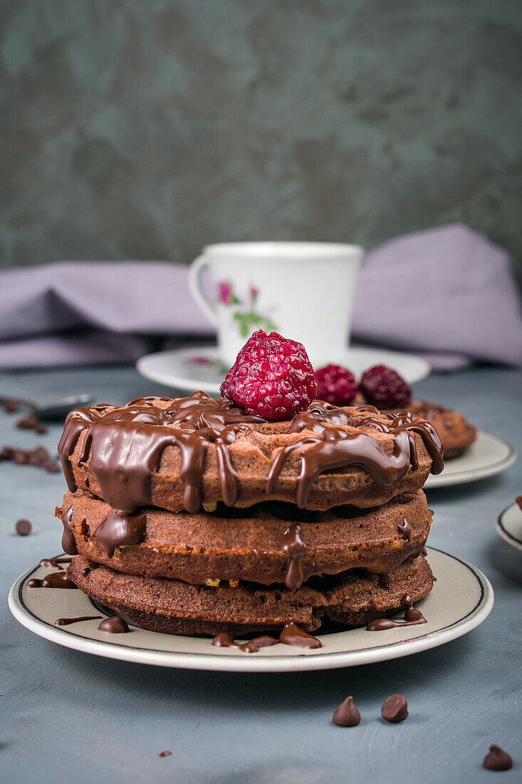 Leckere süße Waffeln mit Schokoladensauce und Himbeeren, serviert auf einem Tisch mit weißer Tasse und Schokoladendrops in einer hellen Küche