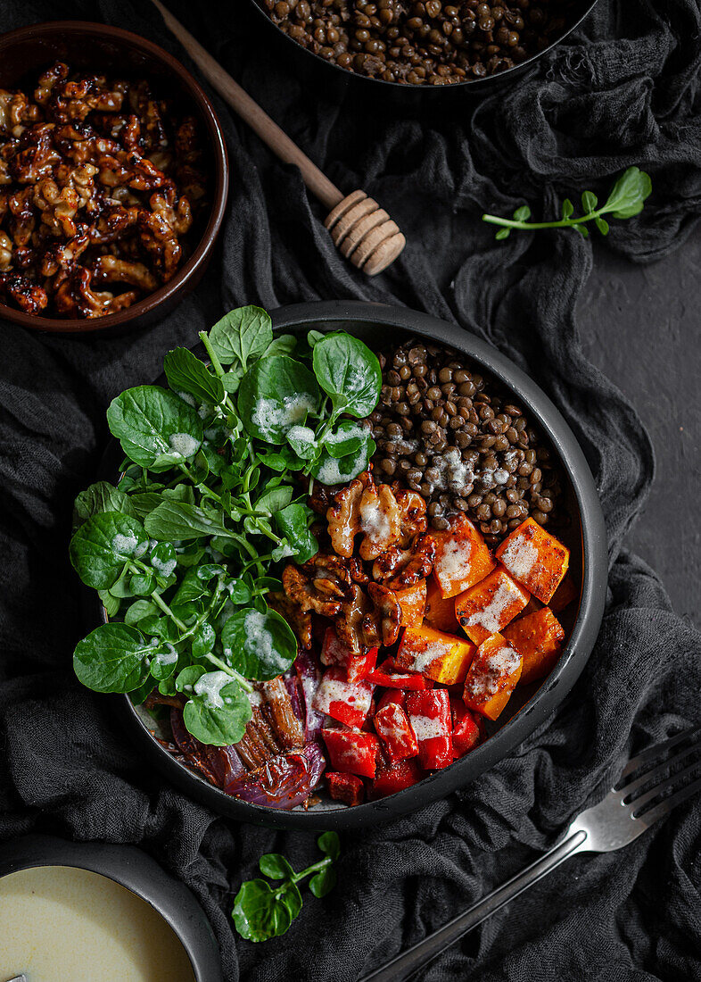 Draufsicht auf Eintopf mit Linsen neben Salat mit Kürbis und Paprika, dekoriert mit grünen Basilikumblättern auf schwarzem Hintergrund neben Salatdressing