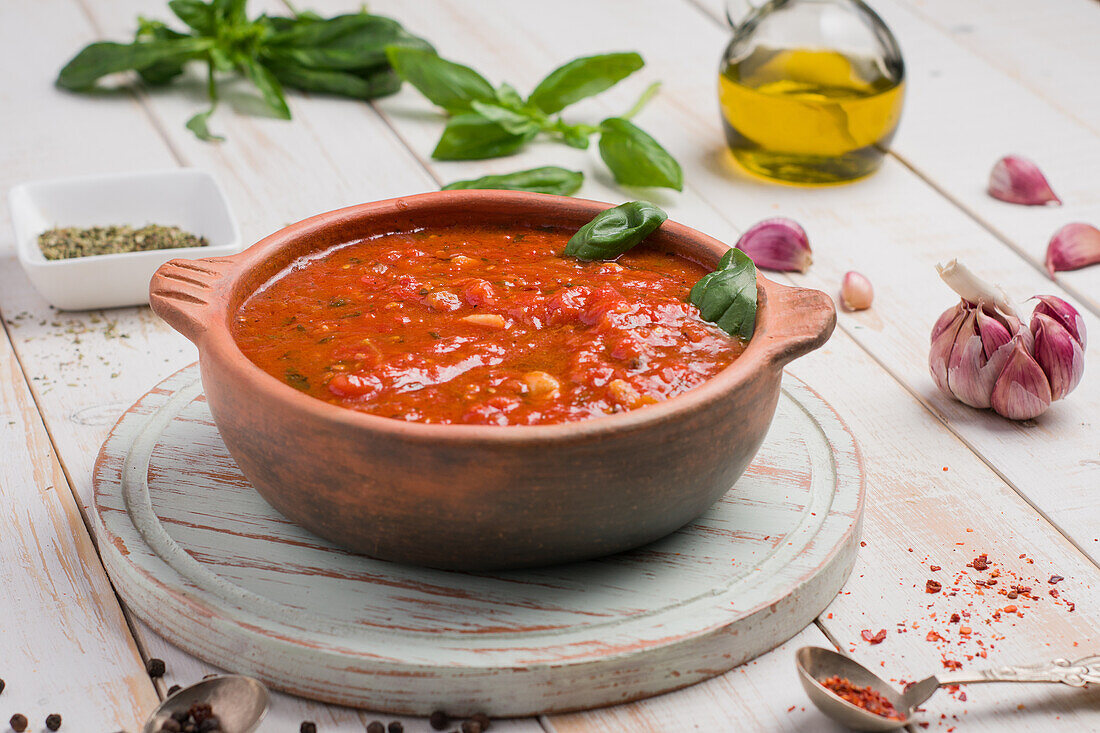 Holzschüssel mit roter Marinara-Soße aus Tomaten und Basilikumblättern auf dem Tisch, dazu Olivenöl und Knoblauch