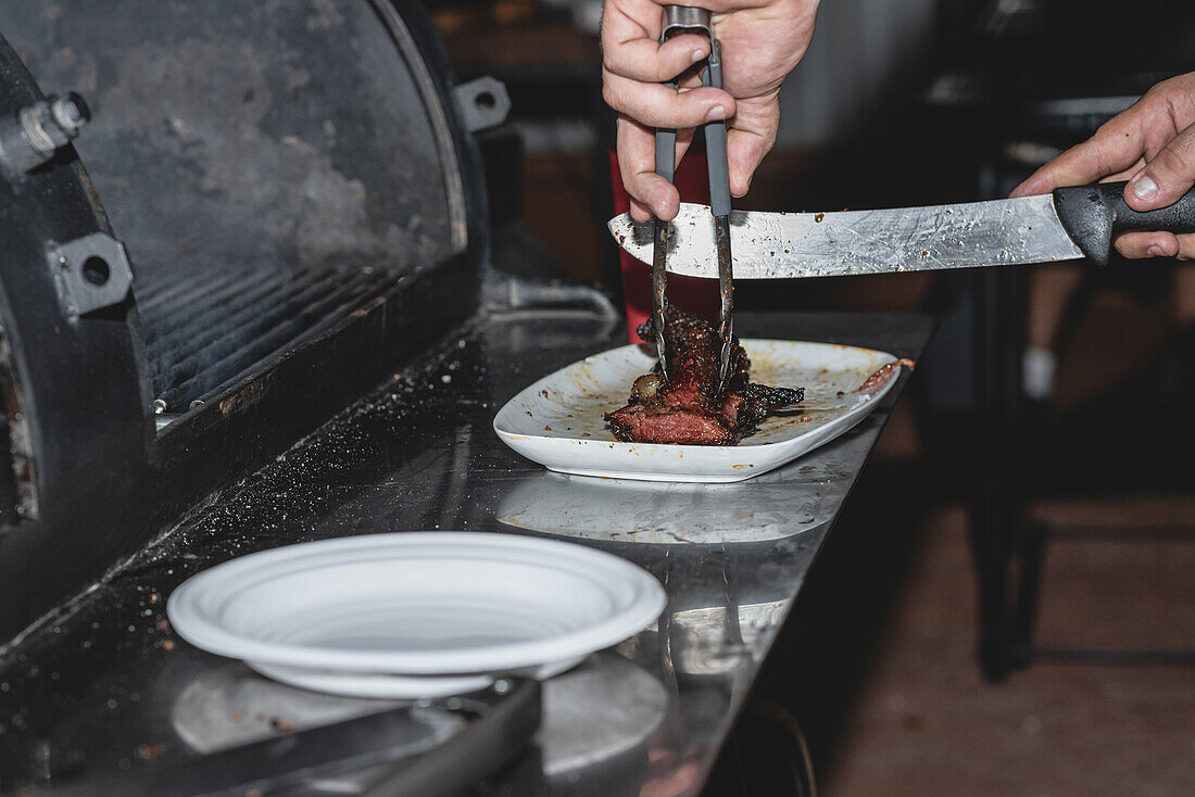 Abgeschnittener gesichtsloser männlicher Koch, der gegrilltes Schweinefleisch mit Zange und Messer schneidet, während er in der Nähe eines modernen Grills in einem Café steht