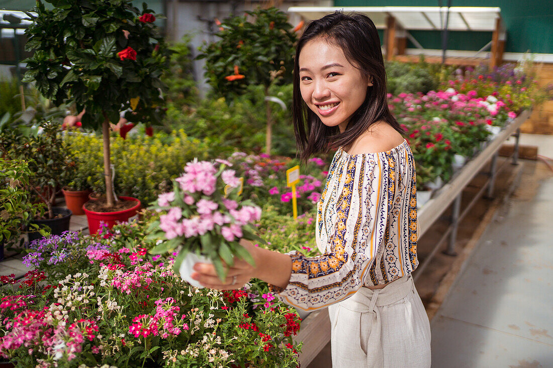 Sincere young ethnic female shopper selecting blooming flowers with pleasant scent in garden shop in daytime