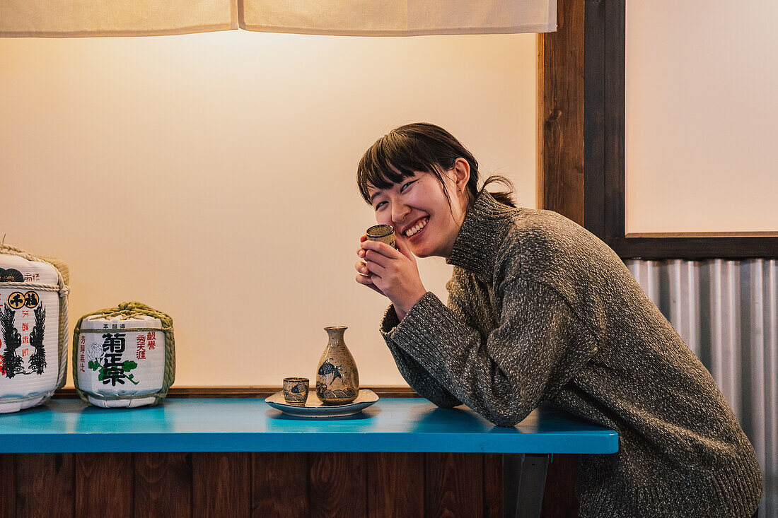 Seitenansicht einer überglücklichen Asiatin im Pullover, die an einem blauen Tisch mit einem traditionellen Getränk in einem Café sitzt