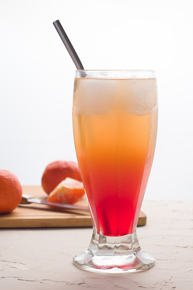 Glass of refreshing Sunrise cocktail with ice cubes and straw served on table with fresh oranges