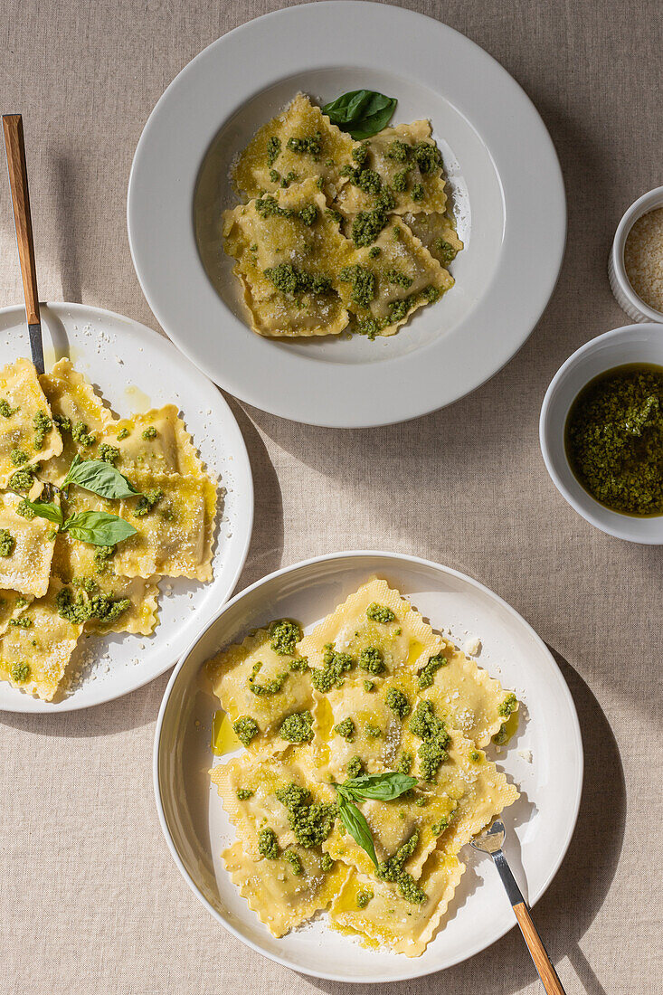 Draufsicht auf appetitlich gekochte Ravioli mit grüner Soße und Kräutern auf weißen Tellern mit Gabeln auf dem Tisch