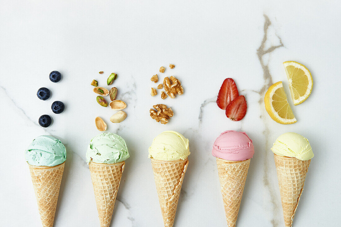 Top view of assorted appetizing ice cream cones with blueberry pistachio walnuts strawberry and lemon composed on marble table with ingredients in kitchen