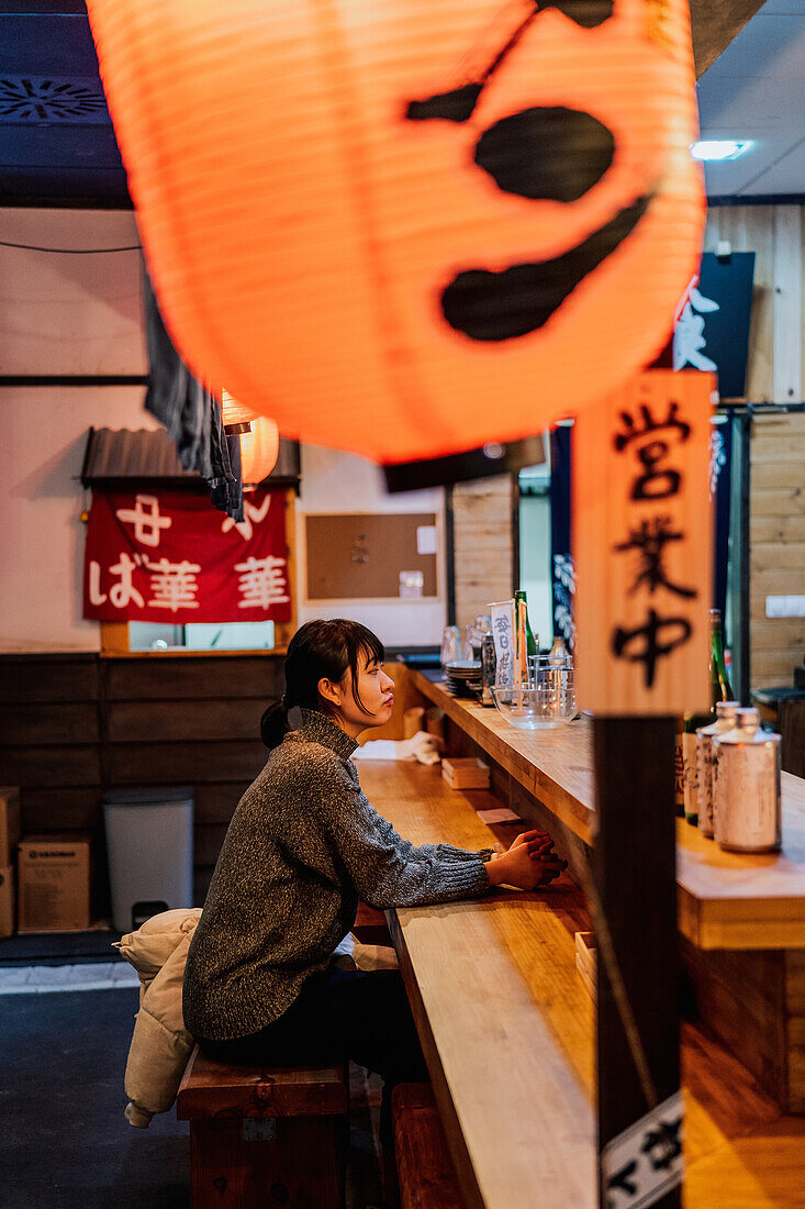 Side view of young Asian woman in casual wear sitting at wooden counter while waiting for order in ramen bar