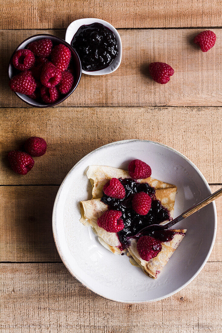 Draufsicht auf leckere Crêpes mit süßer Erdbeermarmelade auf einem Teller neben einem Löffel auf einem Holztisch