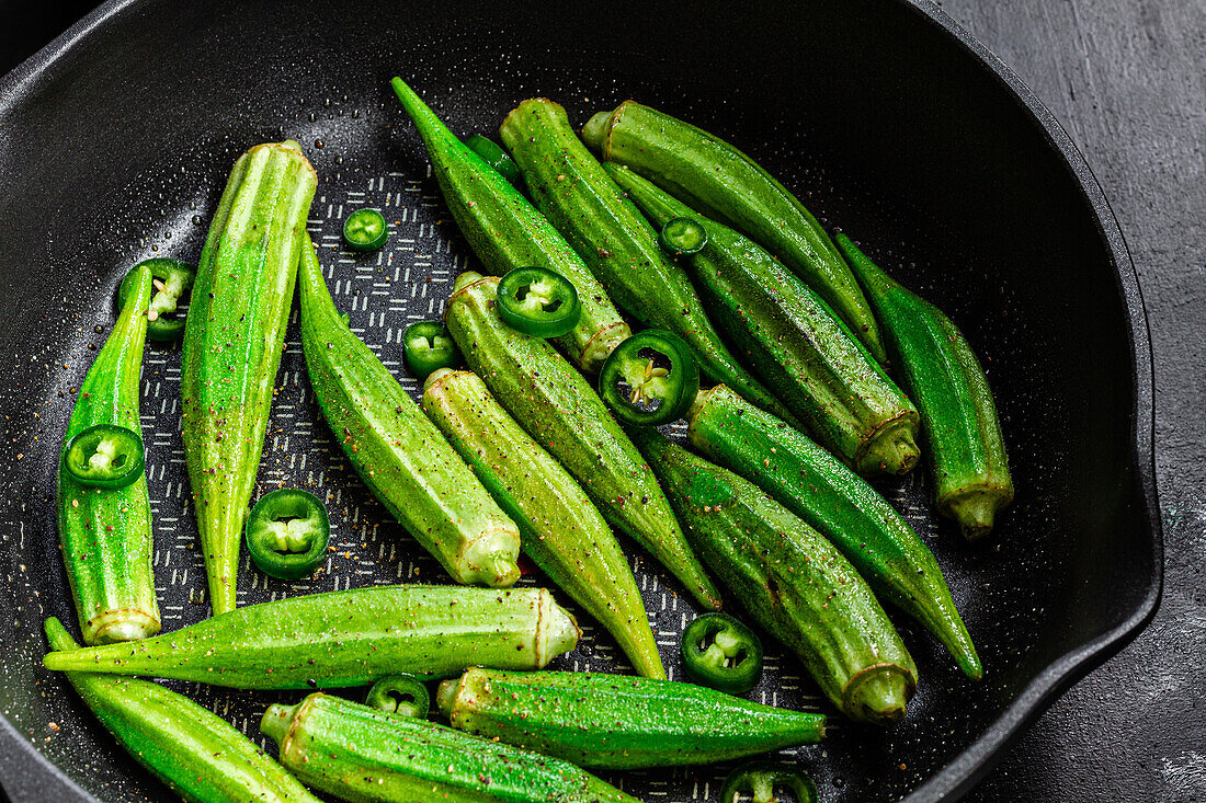 Von oben aufgerissene frische Okra in der Pfanne mit grünem Pfeffer auf dunklem Hintergrund