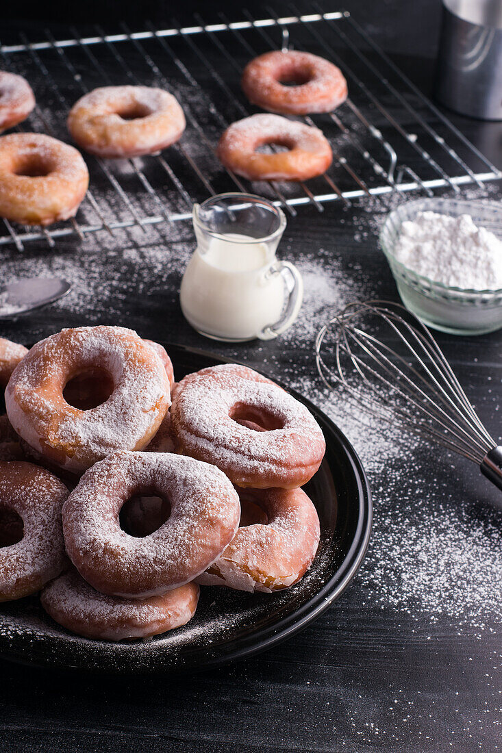 Süße gebratene Krapfen auf einem Teller neben einem Metallkühlregal und einem Milchkännchen auf einem schwarzen, unordentlichen Tisch mit Puderzucker