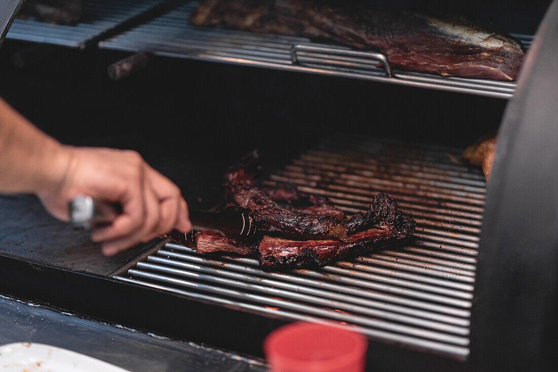 Eine gesichtslose Person mit einer Zange grillt Fleisch auf einem Rost in einem heißen Grill in der Nähe von Soße während des Kochvorgangs