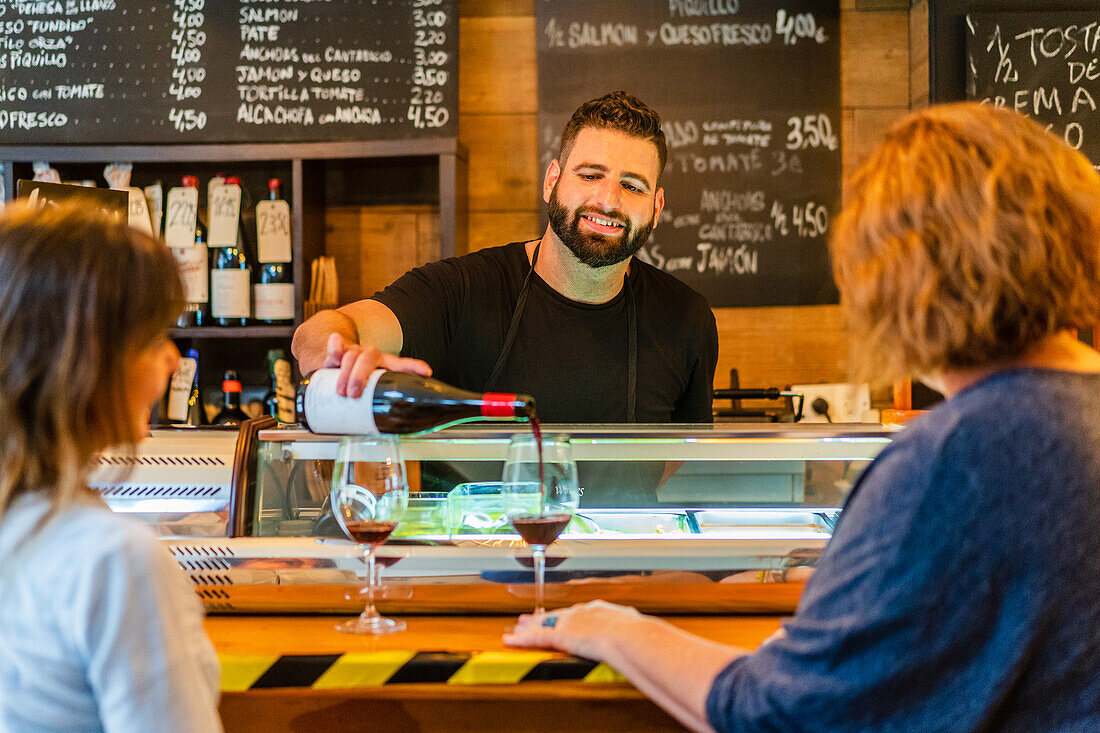 Concentrated barman standing at counter and pouring red wine into glasses for unrecognizable woman customer in cafe