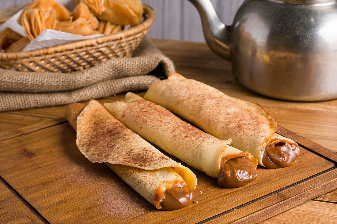 Frisch gerollte Crêpes mit süßer Dulce de Leche-Füllung, serviert auf einem hölzernen Schneidebrett auf einem Tisch mit Wasserkocher in der Küche