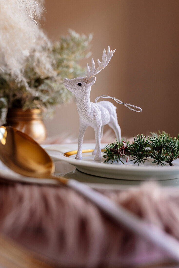 From above christmas table settings with a wreath decoration and white and golden dinnerware and white reindeer on fluffy placemat on pink tablecloth