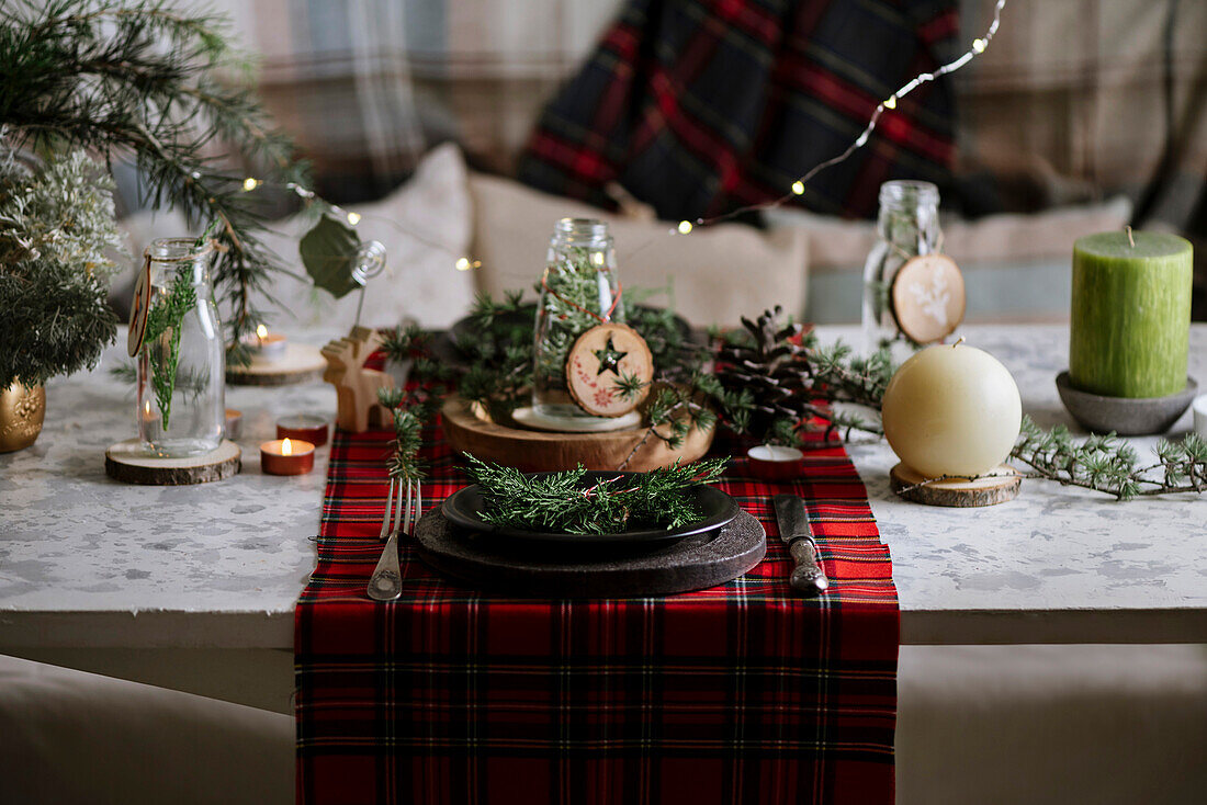 Weihnachtlich gedeckter Tisch mit Kranz auf dem Teller, dekorativem Holzschmuck und rot kariertem Tischtuch mit gelben Lichtern im Hintergrund