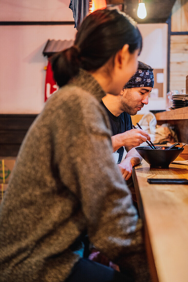 Seitenansicht einer Frau und eines Mannes, die sich beim Essen asiatischer Speisen an einem Holztresen in einem modernen Café unterhalten