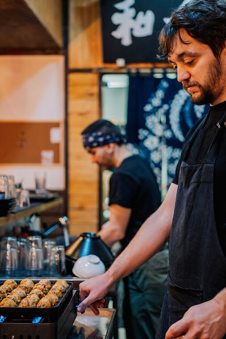 Seitenansicht eines männlichen Kochs in schwarzer Uniform und Kopftuch beim Kochen eines asiatischen Gerichts namens Ramen in einem modernen Café