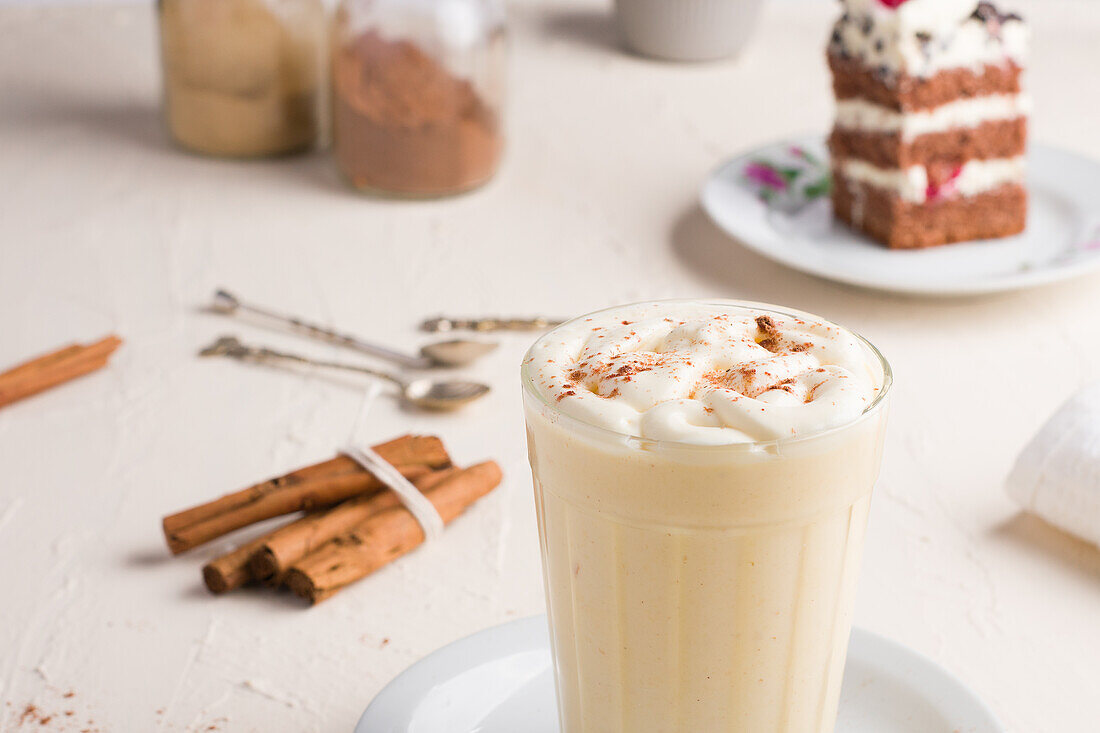 Glas Milchpunsch mit Zimtpulver auf geschlagenem Eiweiß vor Kuchenstück auf Cafeteria-Tisch vor hellem Hintergrund