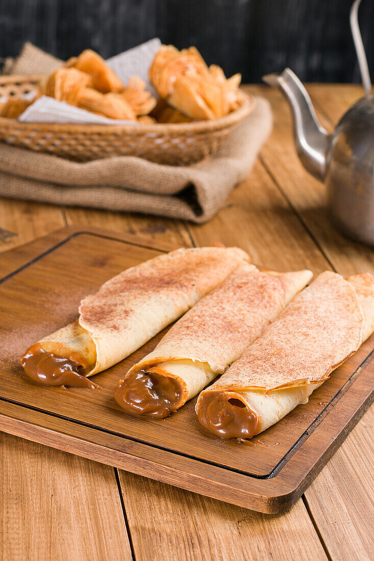 Frisch gerollte Crêpes mit süßer Dulce de Leche-Füllung, serviert auf einem hölzernen Schneidebrett auf einem Tisch mit Wasserkocher in der Küche