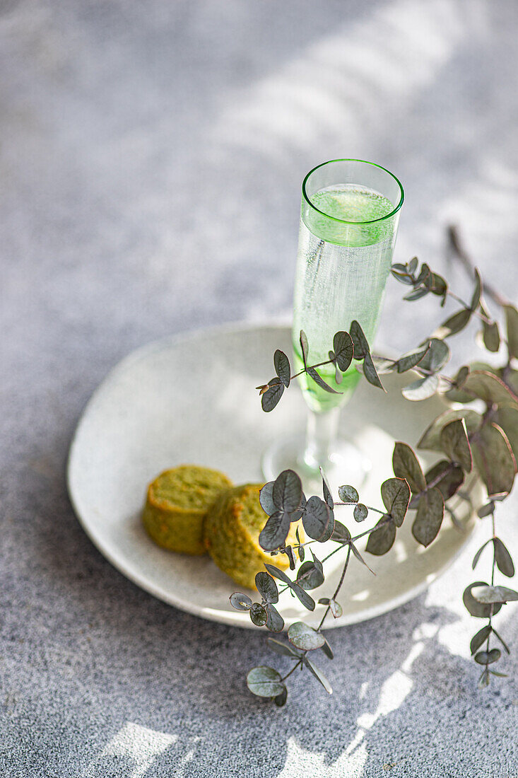 Zarte Eukalyptuszweige akzentuieren ein schlankes Glas Pistazien-Wodka und zwei strukturierte Pistazienkekse auf einem weißen Teller auf einer Betonfläche