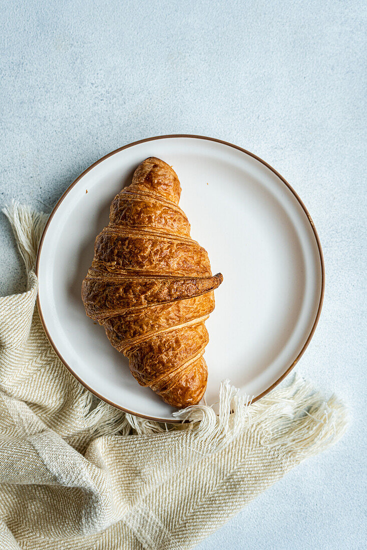 Draufsicht auf ein einzelnes Croissant auf einem runden Teller, das auf einem hessischen Tuch mit strukturiertem Hintergrund ruht und die Einfachheit eines Frühstücks mit Gebäck einfängt