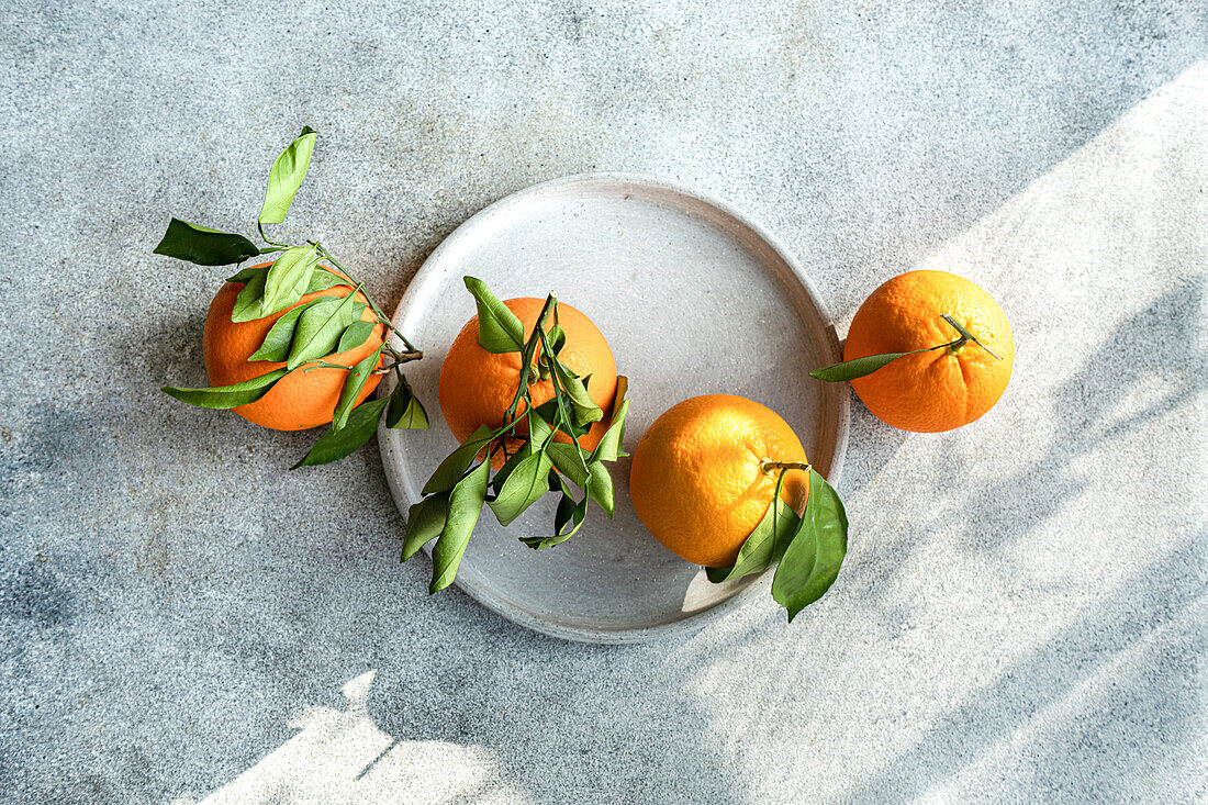 Draufsicht auf ein Trio reifer Orangen, garniert mit üppigen grünen Blättern, präsentiert auf einem grauen runden Steinteller vor einem strukturierten Hintergrund