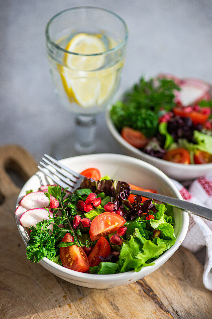 Top view of bowl of fresh vegetable salad with lettuce, arugula, radishes, cherry tomatoes, and pomegranate seeds on a wooden surface, beside a glass of lemon water