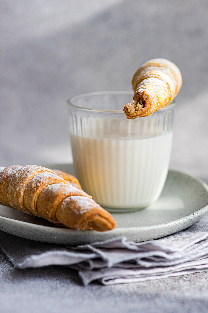 Eine hausgemachte, mit Marmelade gefüllte Gebäcktüte, die in ein Glas Milch getaucht ist, und eine weitere, die auf einem Keramikteller liegt, vor einem strukturierten grauen Hintergrund