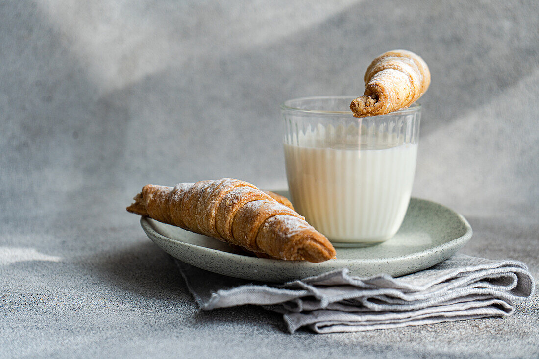 Eine hausgemachte, mit Marmelade gefüllte Gebäcktüte, die in ein Glas Milch getaucht ist, und eine weitere, die auf einem Keramikteller liegt, vor einem strukturierten grauen Hintergrund