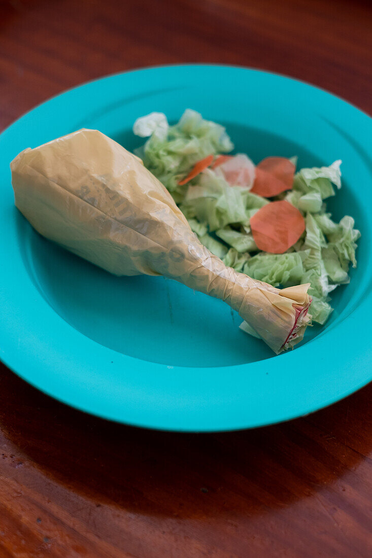 Eine gefälschte Hühnerkeule aus einer verdrehten Plastiktüte, begleitet von einem Salat aus Papierausschnitten auf einem hellblauen Kinderteller.